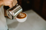Person pouring coffee from a portafilter into a white cup with a latte art design.