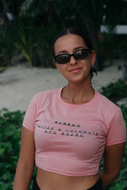 Woman wearing a pink t-shirt with text, standing outdoors with greenery in the background