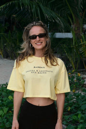 Woman wearing a yellow crop top with text, standing outdoors with greenery in the background