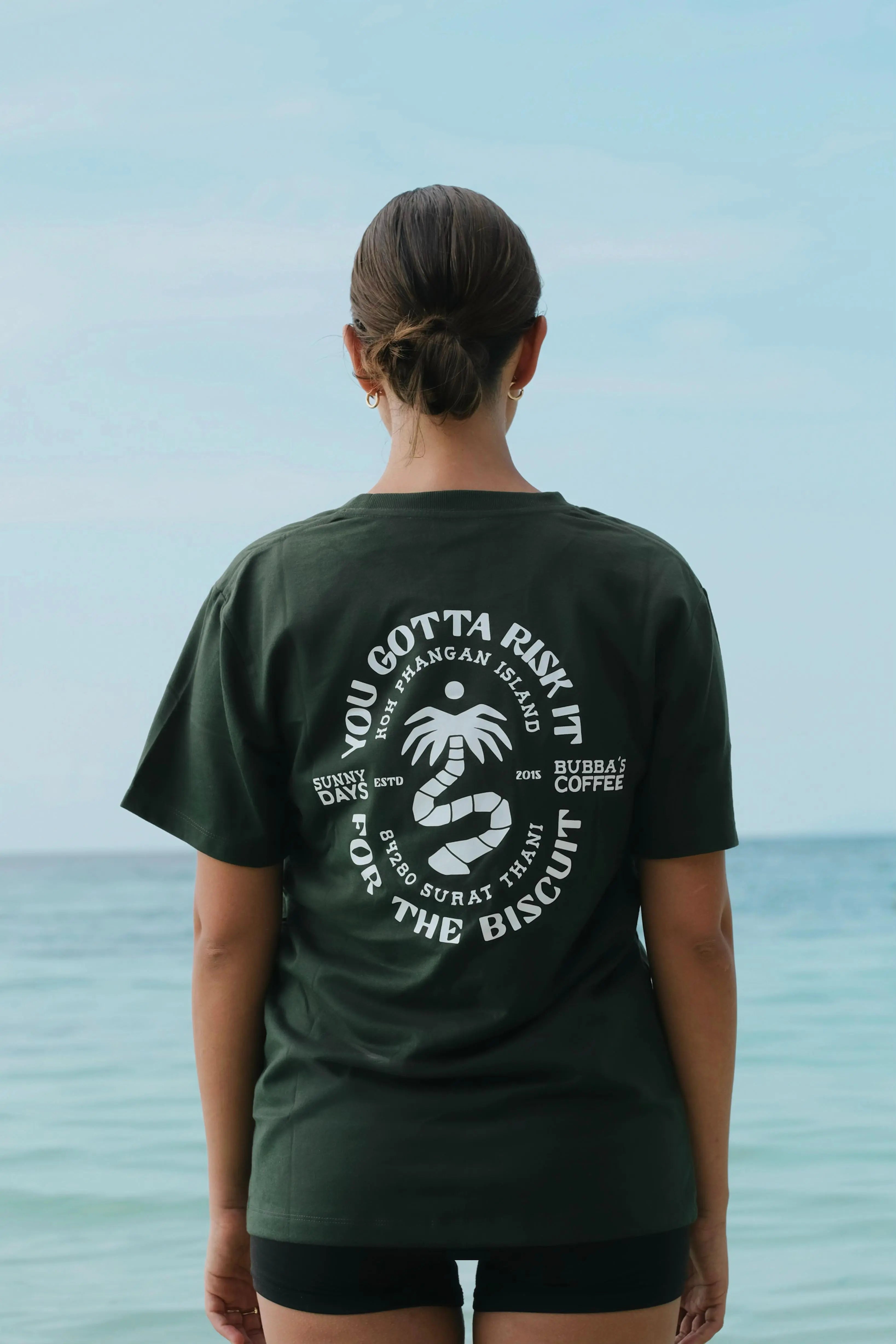 Person wearing a green t-shirt with text and graphics, standing by the ocean.