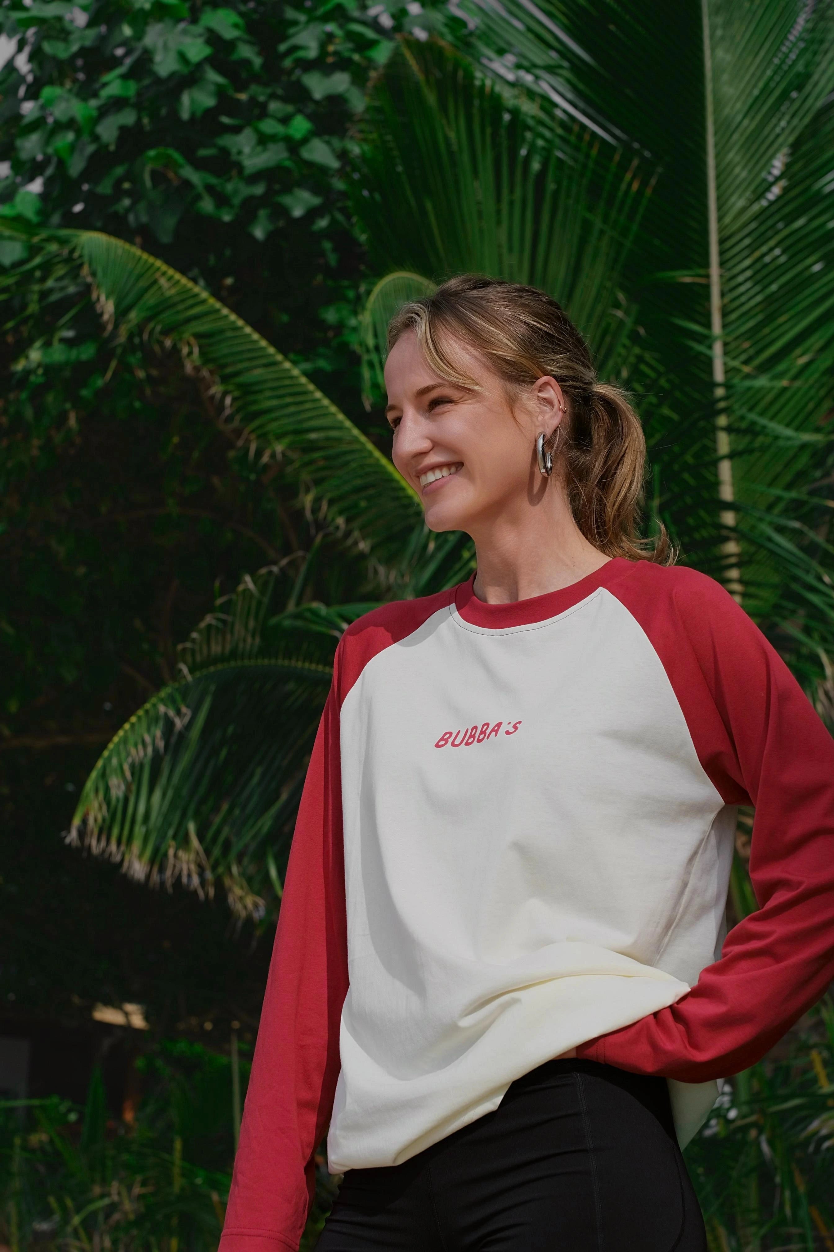 Woman wearing a red and white baseball shirt with 'BUBBA'S' logo, standing in front of green foliage.