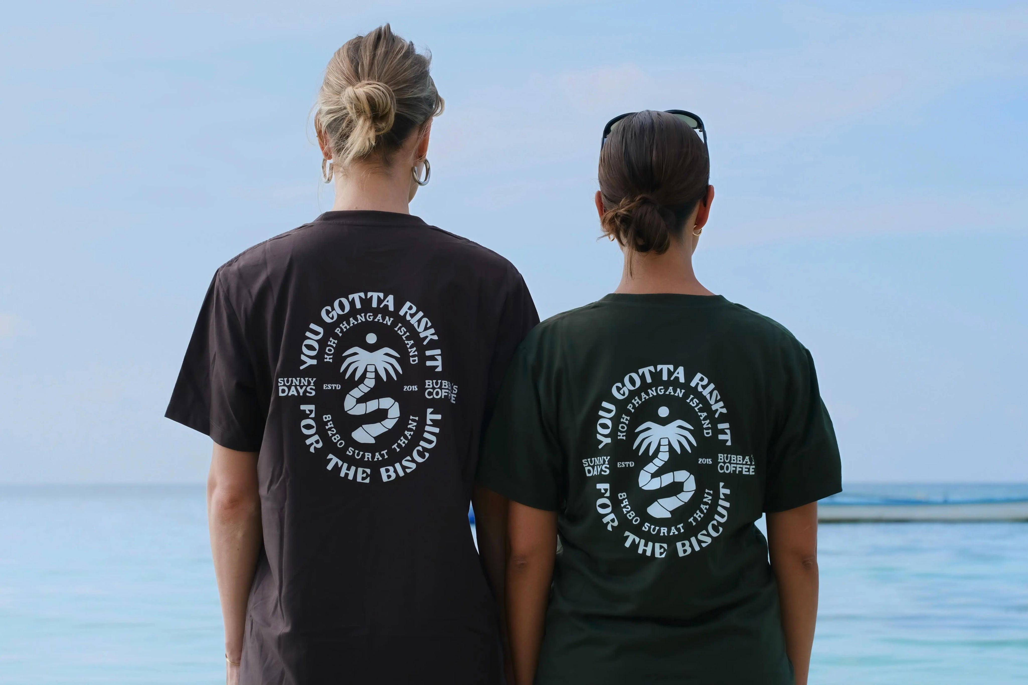 Two people wearing black t-shirts with a palm tree design and text, standing by a body of water.