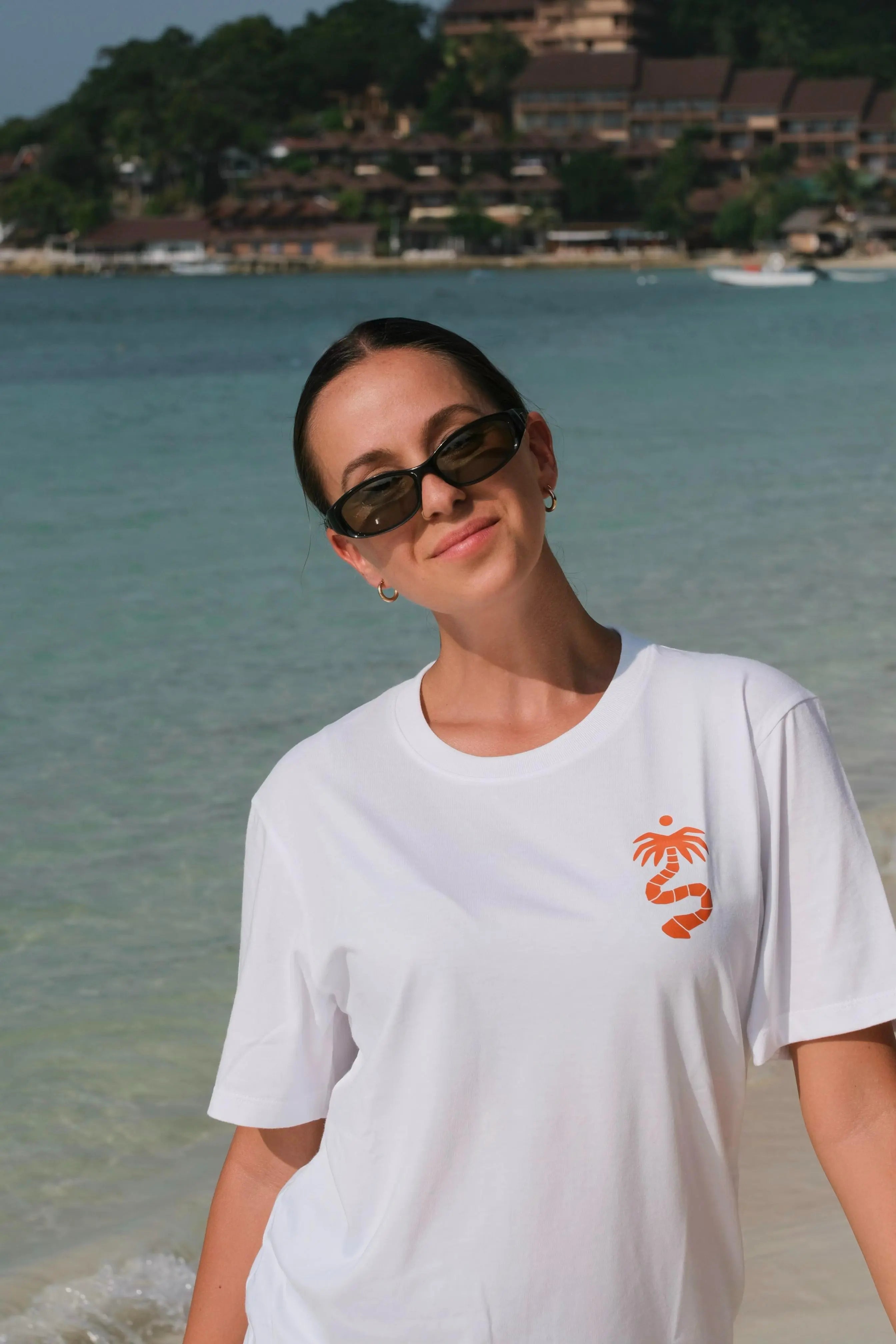 Woman wearing sunglasses and a white t-shirt with a palm tree design on a beach.