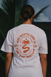 Person wearing a white t-shirt with red text and graphics, standing outdoors with palm trees in the background.