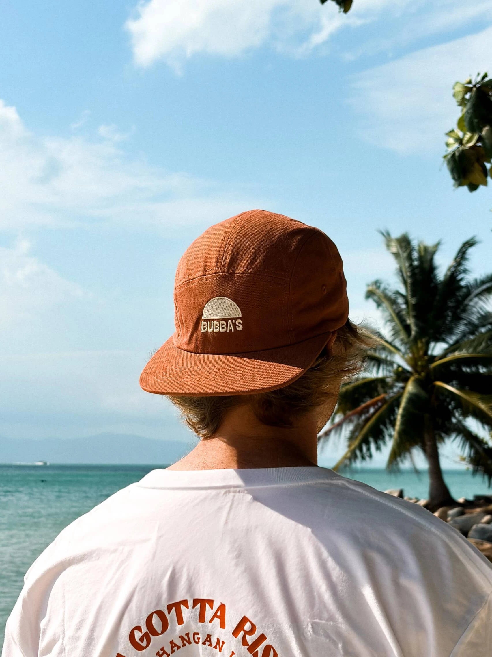 Person wearing a brown cap with a logo, standing on a beach with palm trees and ocean view.