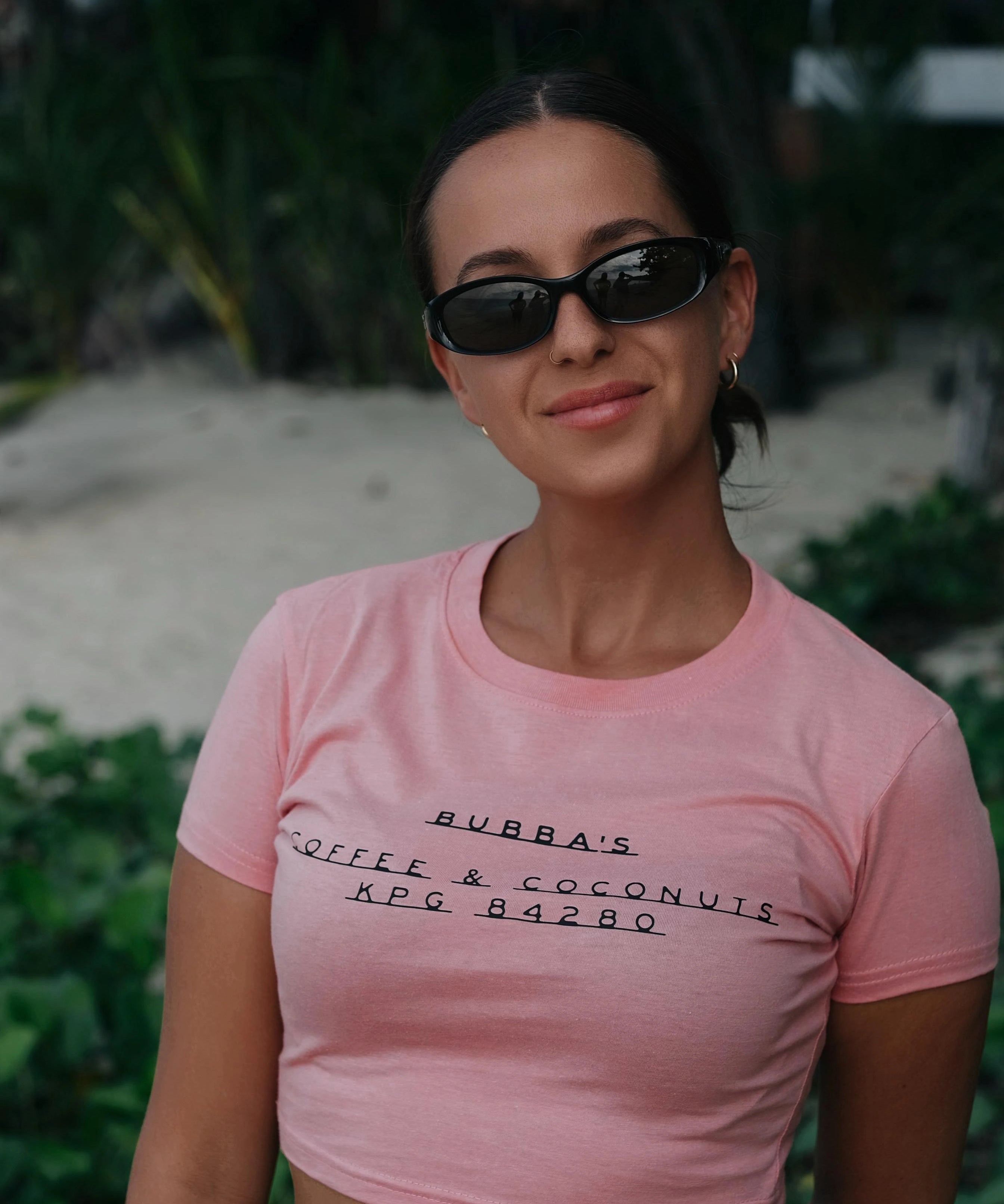 Woman wearing a pink t-shirt with text, standing outdoors with greenery in the background