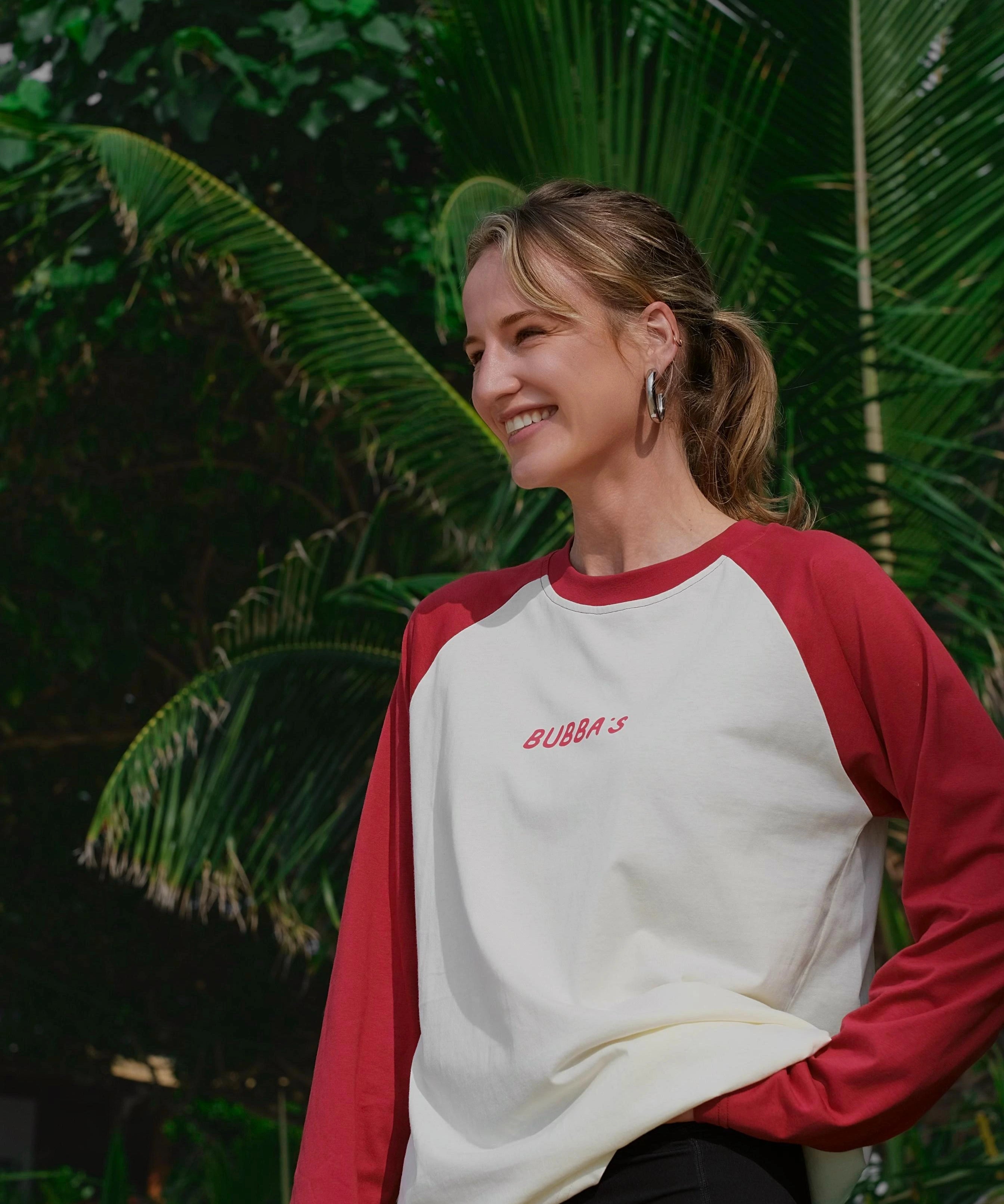 Woman wearing a red and white baseball shirt with 'BUBBA'S' logo, standing in front of green foliage.