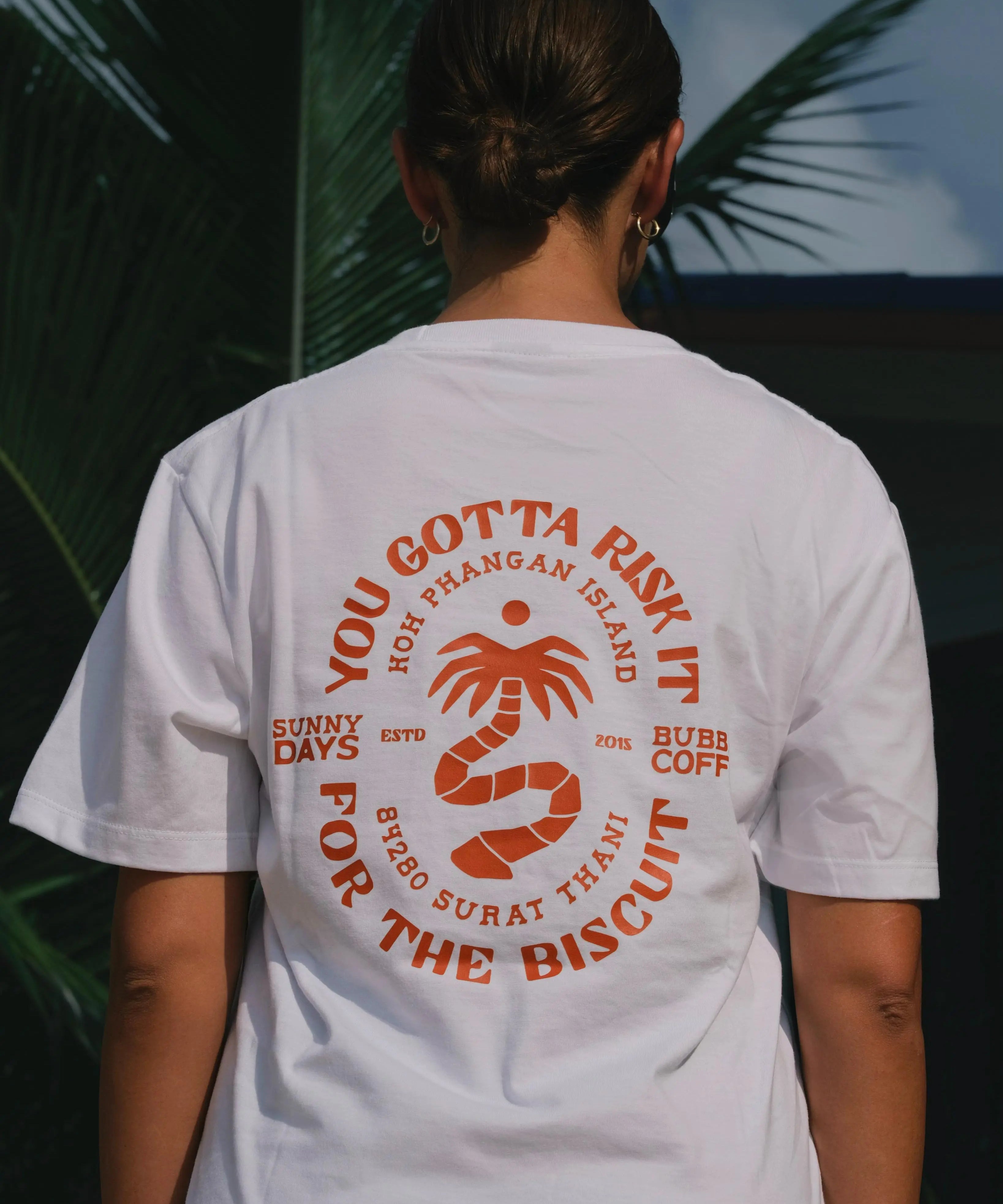 Person wearing a white t-shirt with red text and graphics, standing outdoors with palm trees in the background.
