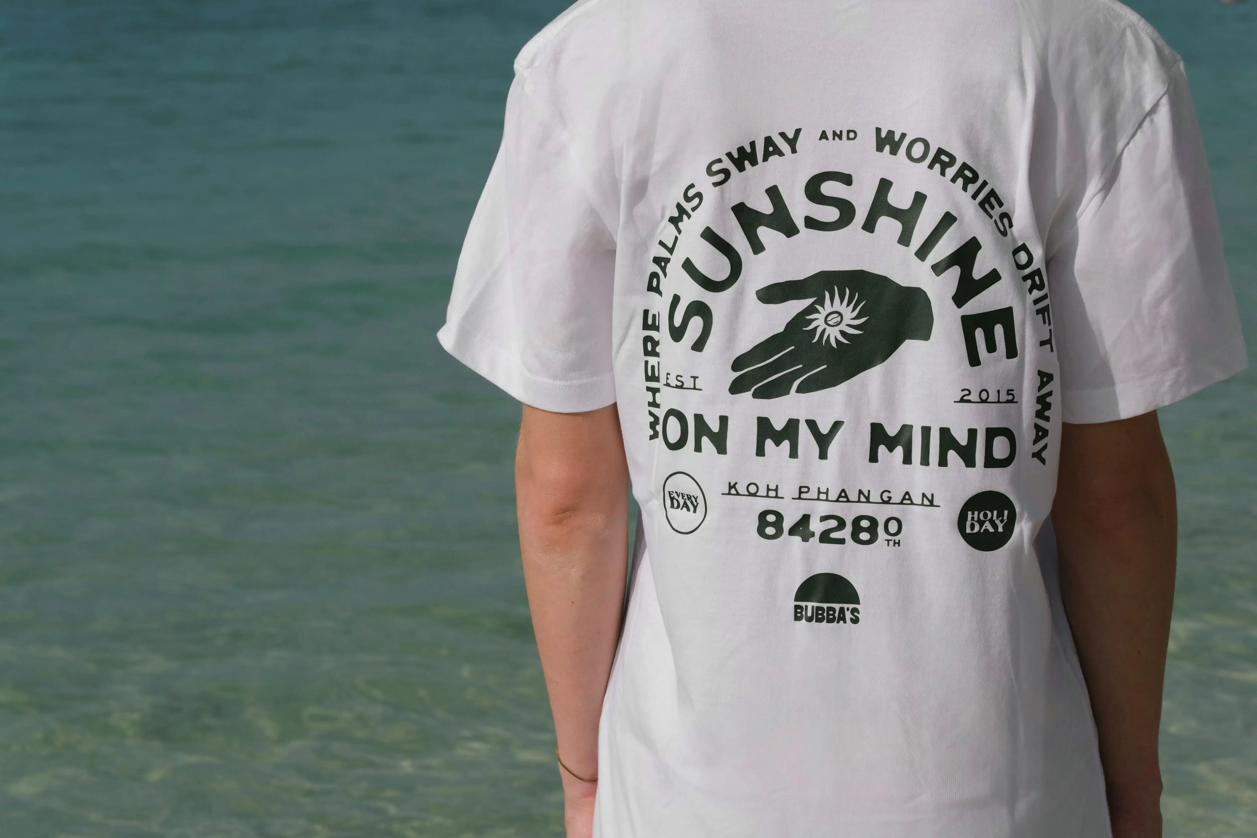 Person wearing a white t-shirt with a black graphic and text on the back, standing by a body of water.