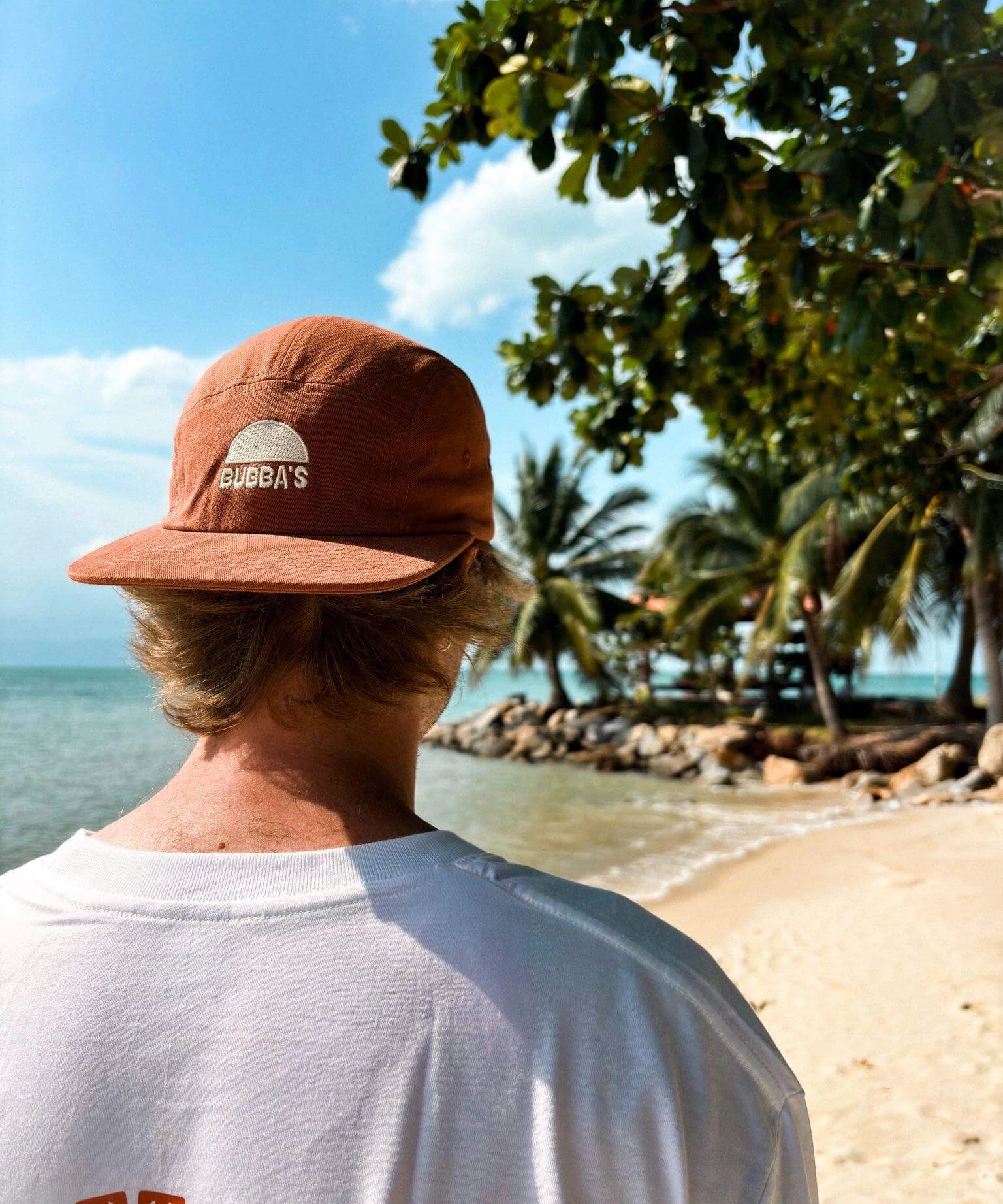 Person wearing a brown cap and white shirt with 'Bugg's' logo, standing on a beach.