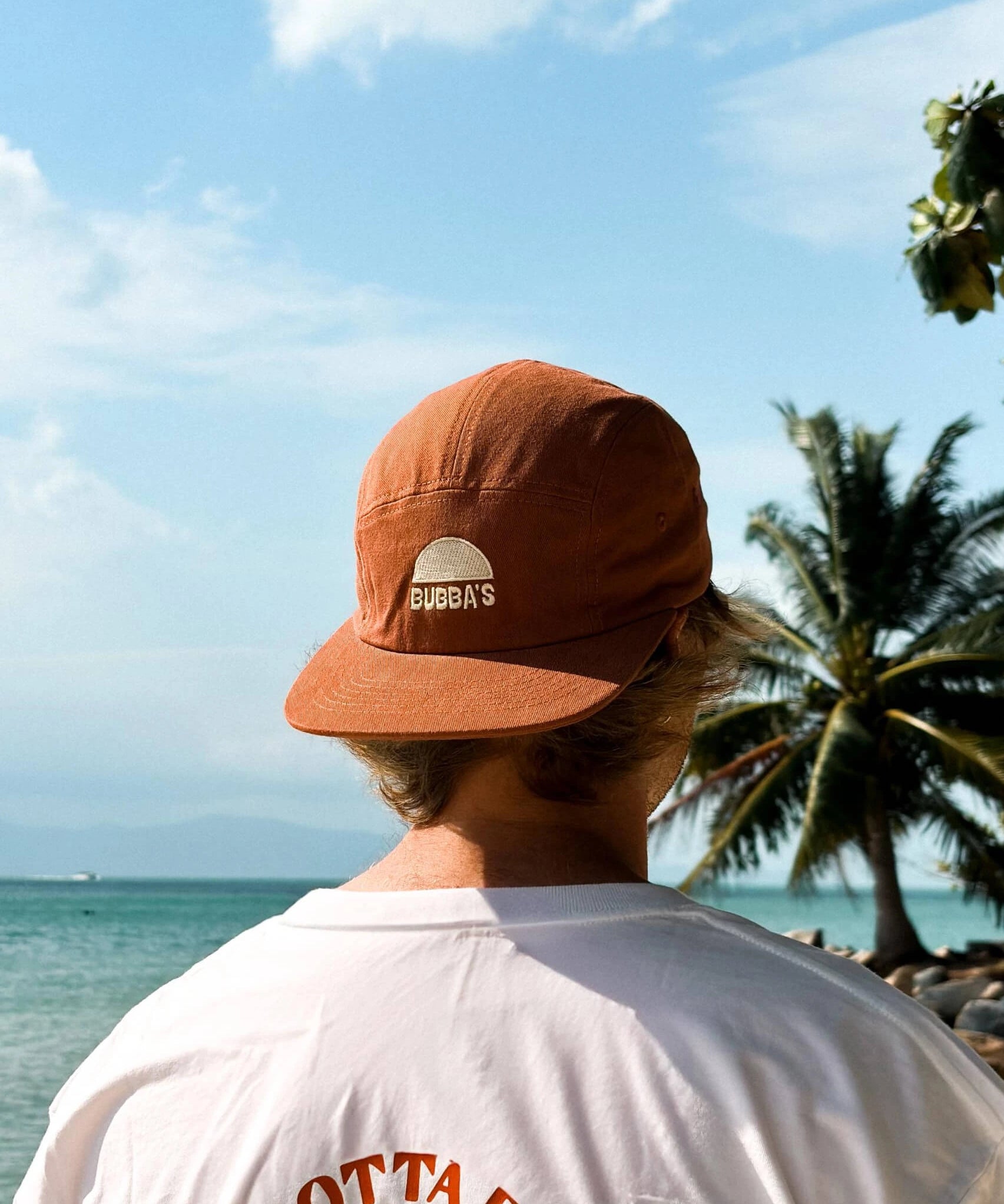 Person wearing a brown cap with a logo, standing on a beach with palm trees and ocean view.